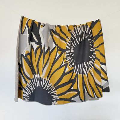 Modern Sunflower Design Cotton Throw hanging on a line, featuring a stylish interpretation of sunflower motifs in light pink, mustard, charcoal grey, and light grey. This 100% cotton throw, handcrafted in the USA, showcases a unique blend of traditional floral charm and modern design.