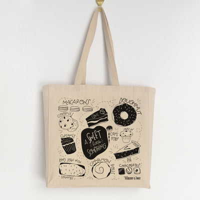 A canvas tote bag with colorful illustrations of sweets and baked goods including doughnuts, chocolates, cake, pie, cupcake, cookies, and macarons, as well as various sprinkles. The text on the bag reads "A Little Something Sweet." Ideal for bakers and fans of baking shows like Great British Bakeoff.