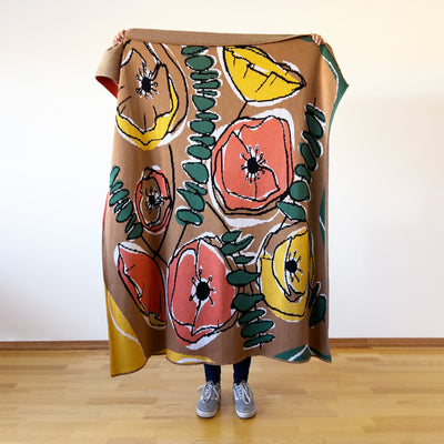 Person holding retro poppy designer cotton throw showing camel brown and hunter green botanical illustration with printmaking-inspired coloring.