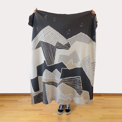 Person holding a large black, white, gray, and tan patterned blanket, making a mountain and starry sky landscape, against a white wall.
