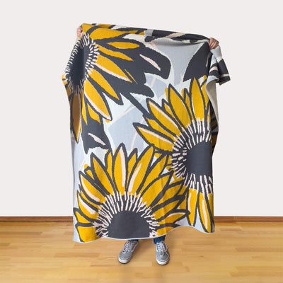 Person holding a sunflower-patterned blanket in a room with wooden flooring.
