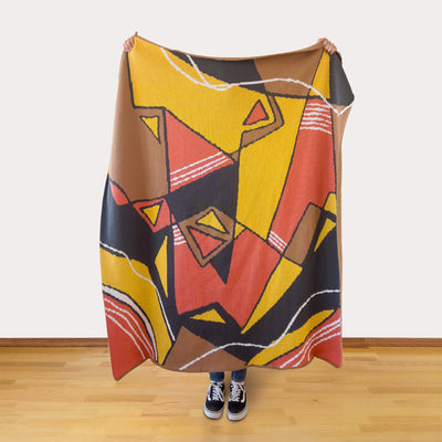 Person holding a colorful abstract patterned blanket, made of yellows, oranges, browns, and grays, on a wooden floor.