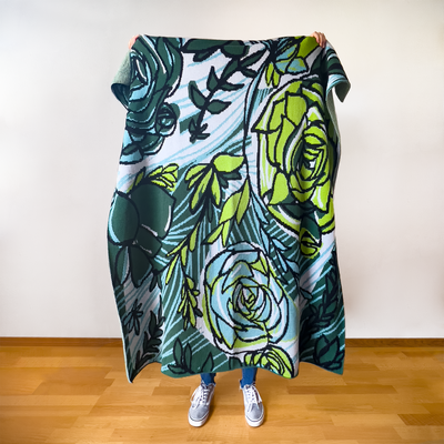 Person in Vans standing on a wooden floor against a white wall, holding a Succulent Inspired Cotton Throw with vibrant turquoise, black, parrot green, hunter green, and gray colors, designed by Tuxberry & Whit, 100% cotton, eco-friendly, crafted in USA.