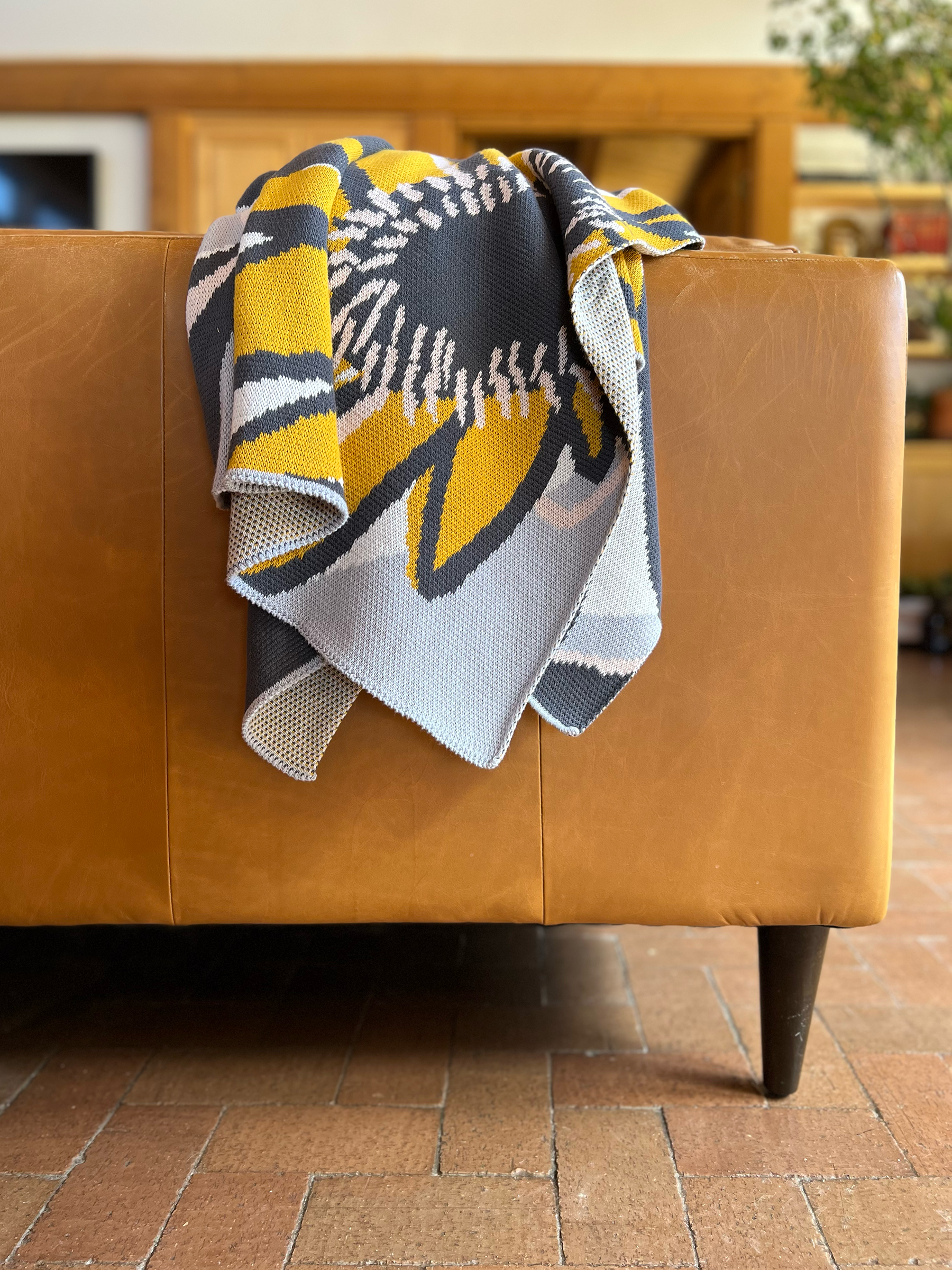 Modern Sunflower Design Cotton Throw elegantly draped over a mid-century modern camel-colored leather couch, creating a stylish contrast. The throw features a unique sunflower pattern in shades of light pink, mustard, charcoal grey, and light grey, beautifully complementing the warm tones of the leather. Crafted from 100% cotton in the USA.