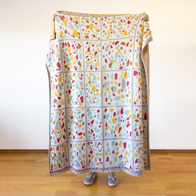 Person holding mid-century terrazzo designer cotton throw showing full coral yellow gray camel aqua speckle pattern on cream background, USA-made