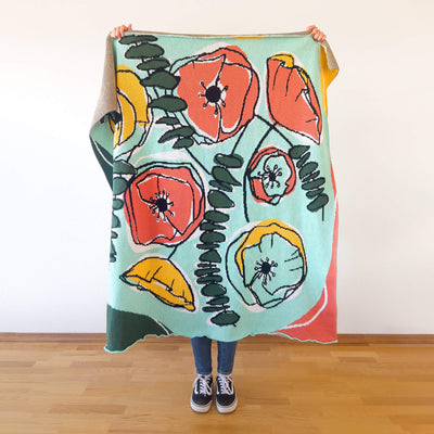 Person holding brightly colored high quality knitted throw with abstract drawing of poppies and eucalyptus.