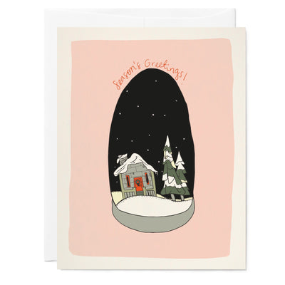 Illustrated holiday greeting card with drawing of  snow globe village scene with little house, trees, and snow falling on a pink background.